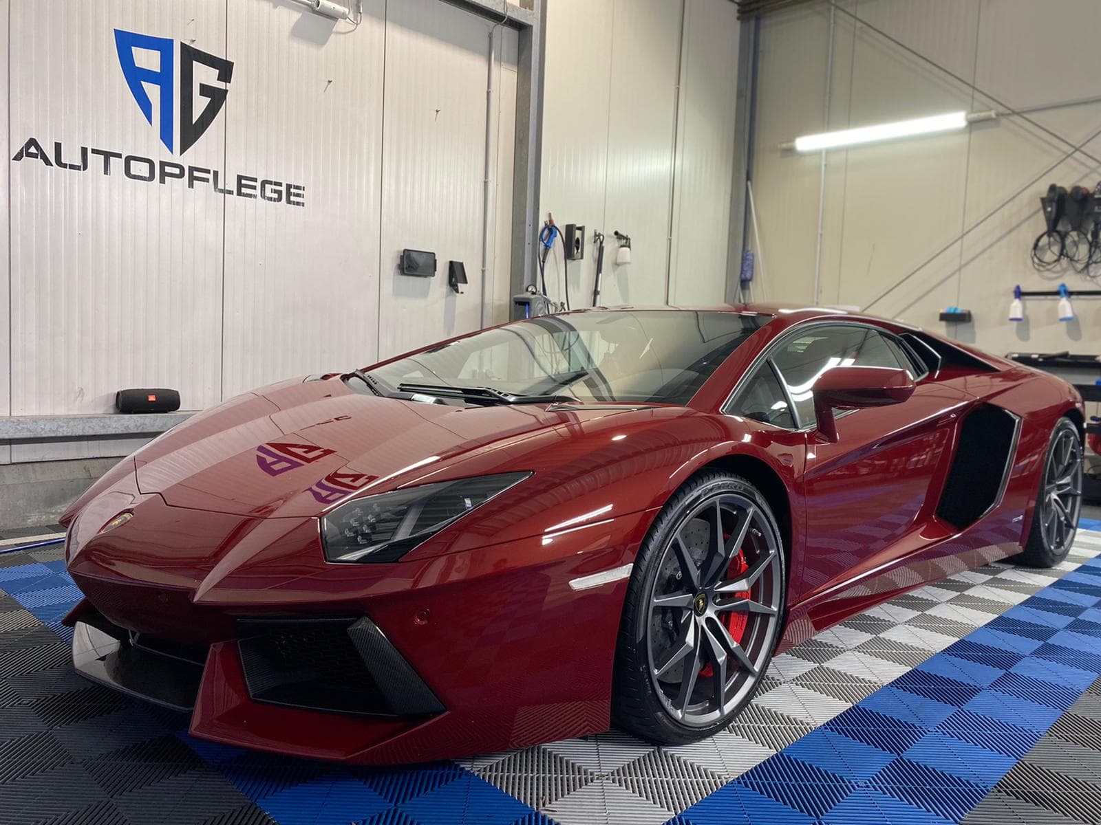 Roter Lamborghini Aventador von schräg vorn in einer hellen Halle mit dem AG Autopflege Logo an der Wand